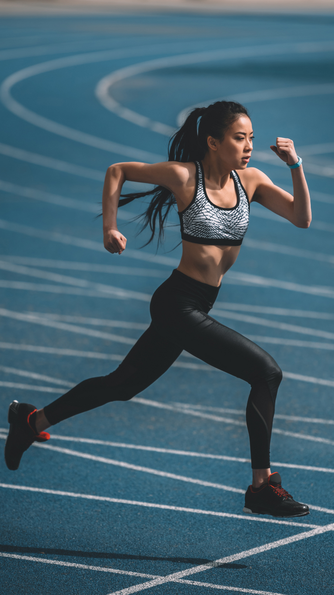 A woman running on a blue athletic track wearing a black and gray patterned sports bra, black leggings, and black running shoes. Elastomers used in sporting industry for activewear and shoes.